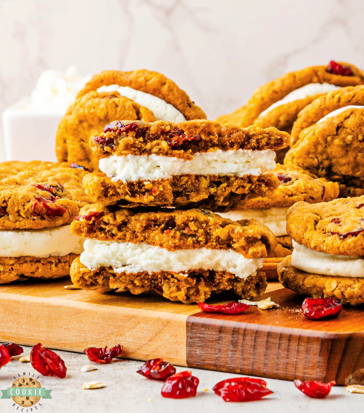 Soft pumpkin oatmeal cookies with dried cranberries and creamy frosting filling stacked on a wooden cutting board.
